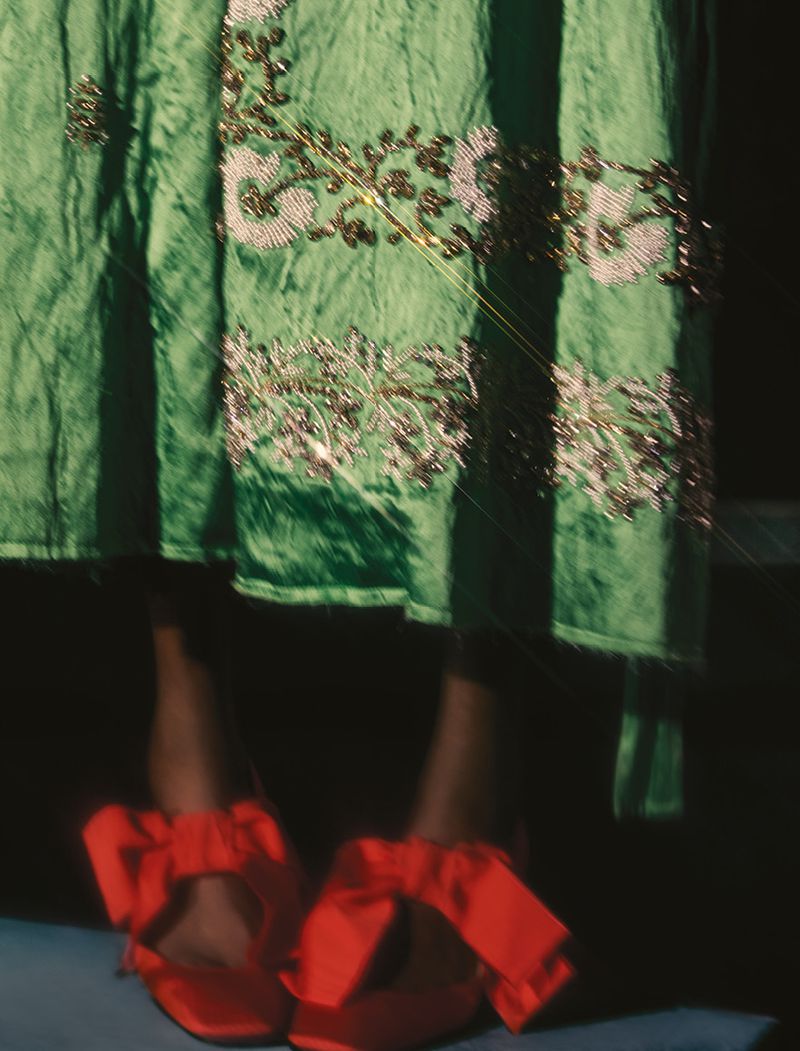 Close-up of a model wearing red ribbon-tie shoes and a green embellished dress, with dark hair, captured in soft, diffused light.