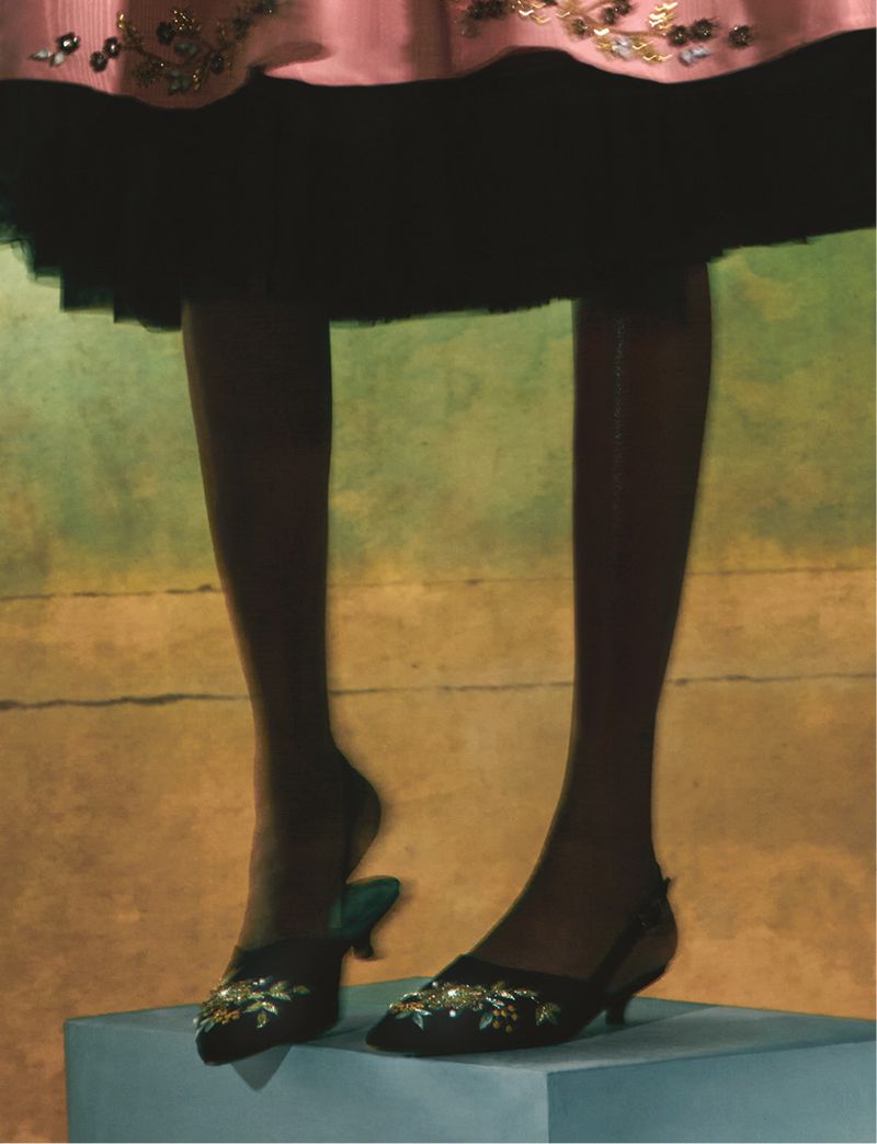 Dark-skinned female legs and feet are showcased, emphasizing elegant black heels adorned with gold floral embellishments. 
