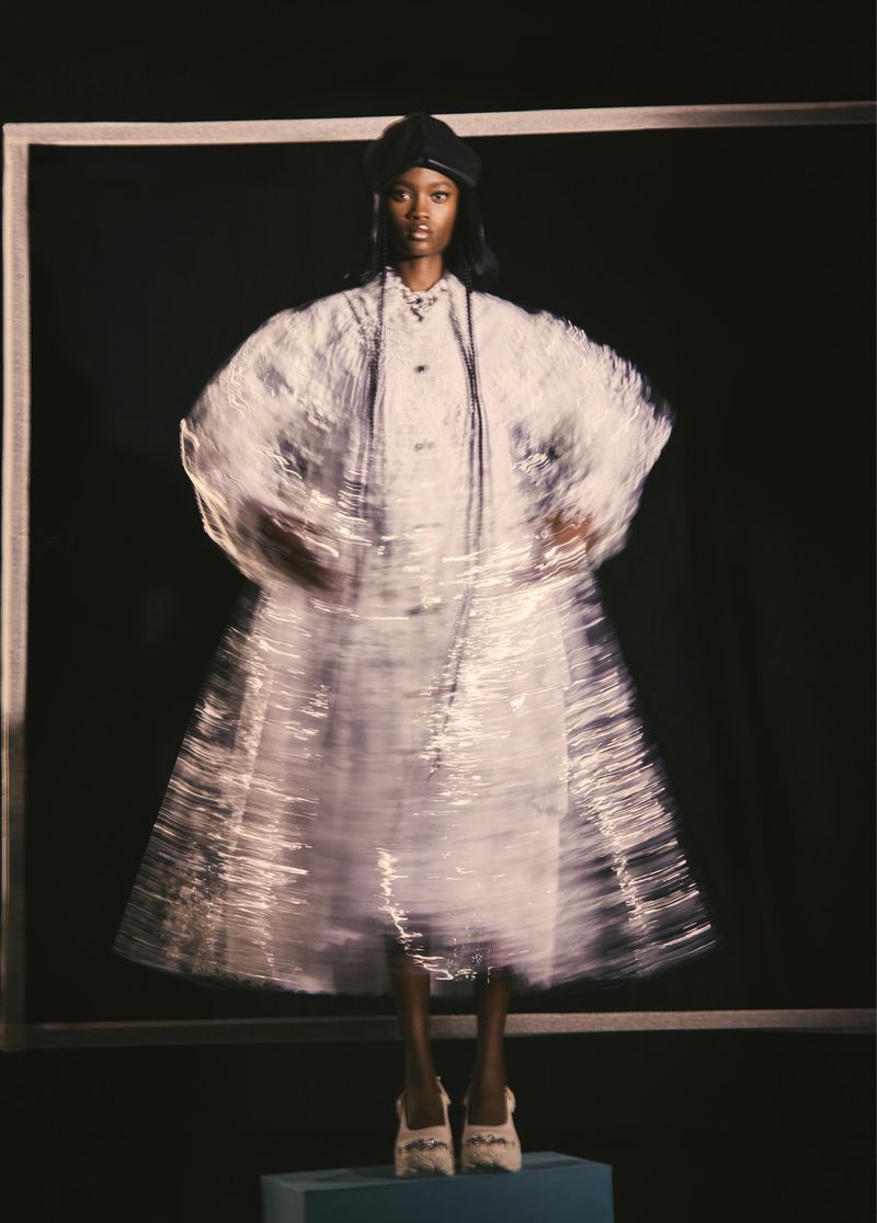 A Black female fashion model poses in a studio wearing a layered, shiny white dress and a black cap against a dark background.