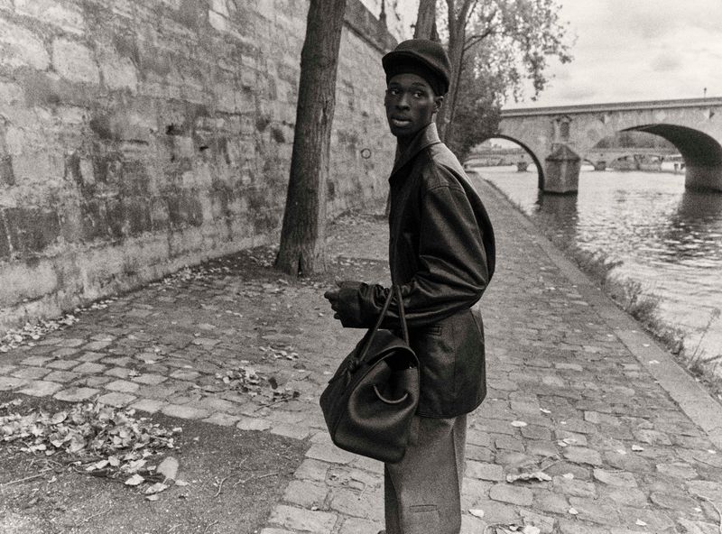 Monochrome portrait of a stylish African male in hat with bag walking along the Seine River in Paris with a stone bridge in background.