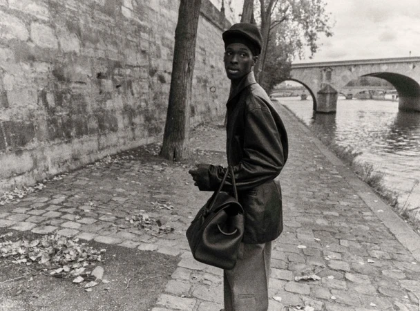 Monochrome portrait of a stylish African male in hat with bag walking along the Seine River in Paris with a stone bridge in background.