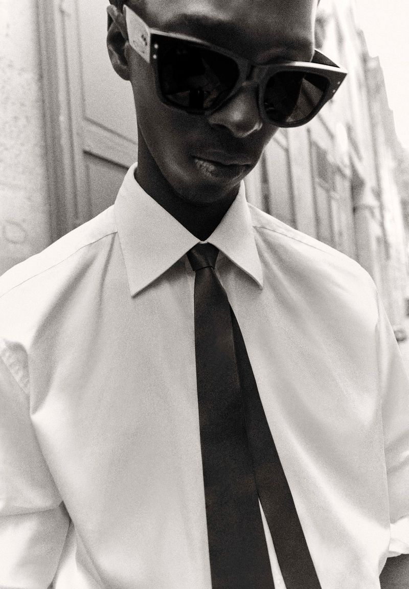 Black and white portrait of a Black man in a dress shirt and tie wearing sunglasses in an outdoor setting with architectural elements.