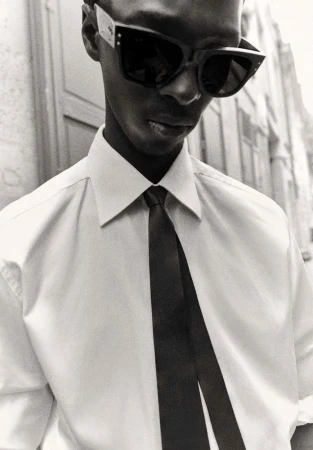 Black and white portrait of a Black man in a dress shirt and tie wearing sunglasses in an outdoor setting with architectural elements.