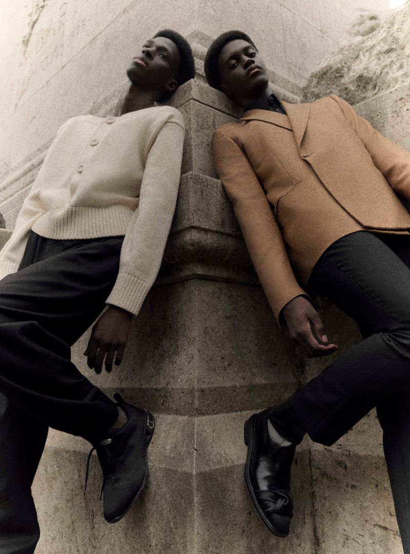 Two dark-skinned young men in fashionable attire lean against a stone pillar in an urban setting, showcasing elegant and vintage style.