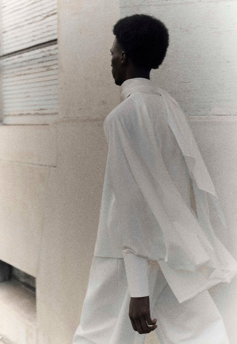 An adult male with afro hairstyle wears white shirt and trousers, walking next to a light beige wall, photographed in soft diffused light.