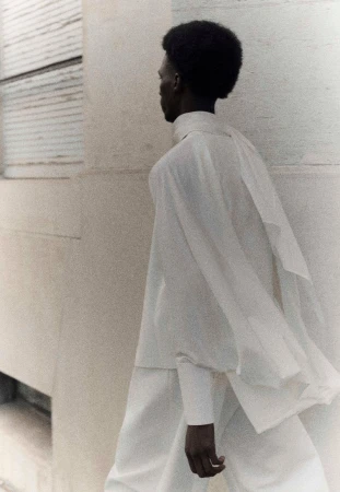 An adult male with afro hairstyle wears white shirt and trousers, walking next to a light beige wall, photographed in soft diffused light.