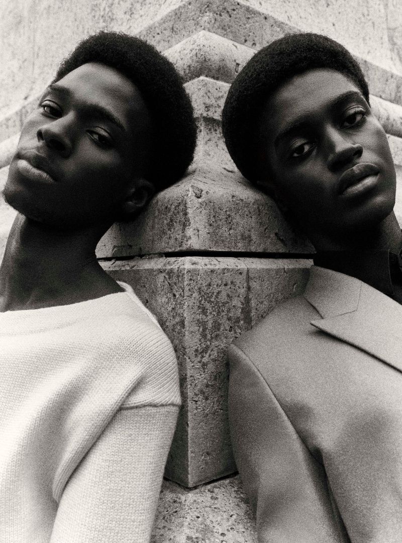 Monochrome portrait of two dark-skinned adult men in a close-up studio shot with a stone structure background, gazing at the camera.