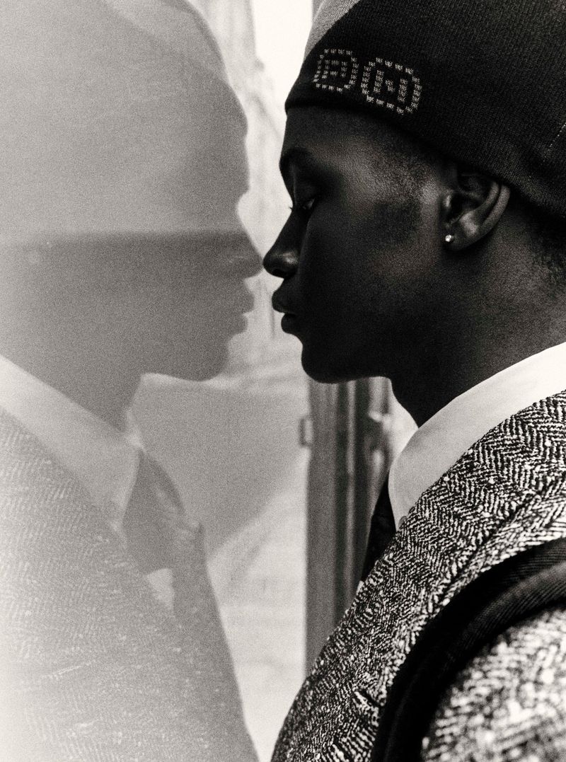 Monochromatic side portrait of a young Black man in a cap and textured jacket, gazing at his blurred reflection in a window.