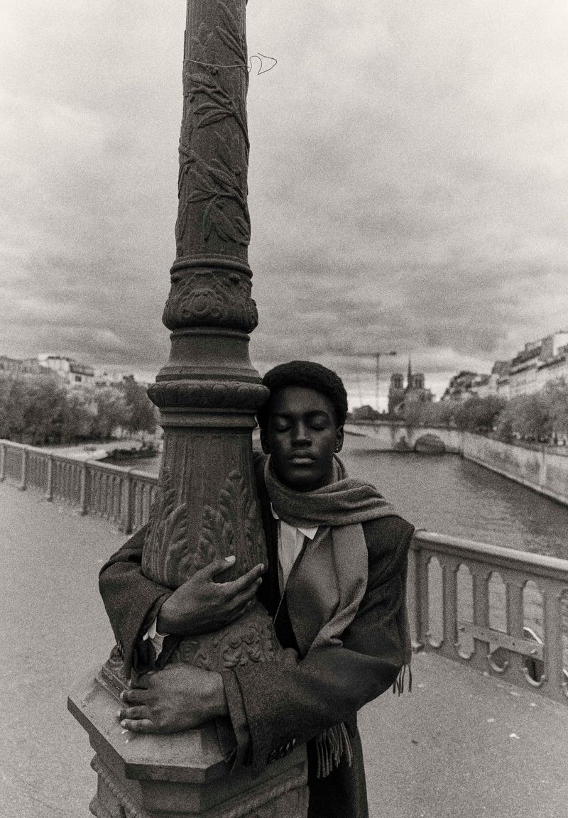 Monochrome portrait of an African American man hugging an ornate lamppost on a bridge with a river and buildings in the background.
