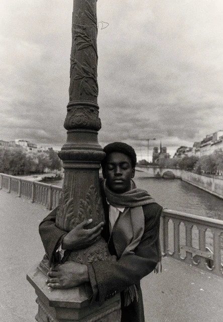 Monochrome portrait of an African American man hugging an ornate lamppost on a bridge with a river and buildings in the background.