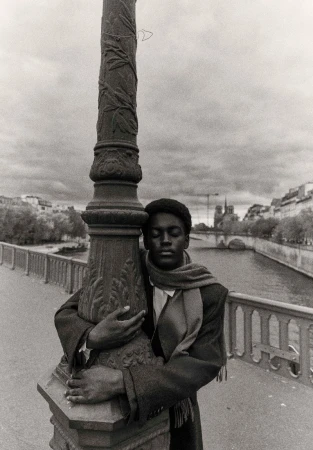 Monochrome portrait of an African American man hugging an ornate lamppost on a bridge with a river and buildings in the background.