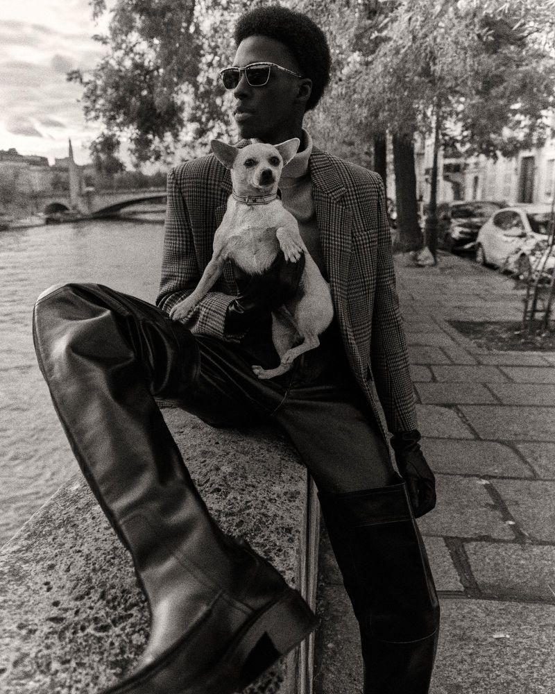 Monochrome portrait of a Black man in sunglasses sitting on a ledge in an urban setting, holding a small dog and wearing high leather boots.