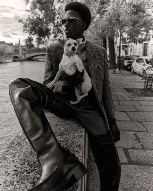 Monochrome portrait of a Black man in sunglasses sitting on a ledge in an urban setting, holding a small dog and wearing high leather boots.