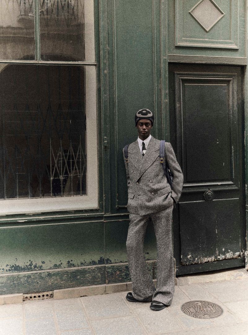 Young man in a grey tweed suit and cap stands against a green wall with a window and a door in an urban outdoor setting.