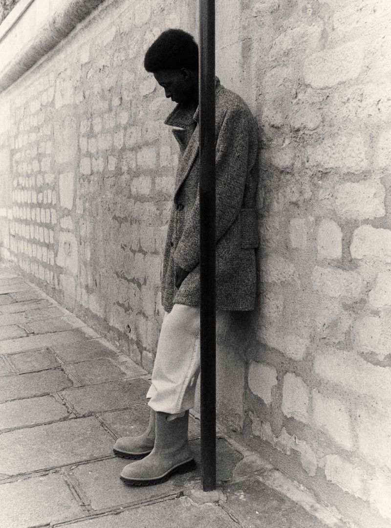Monochrome shot of an adult male standing in an urban setting wearing a coat, trousers, and boots, leaning against a brick wall.