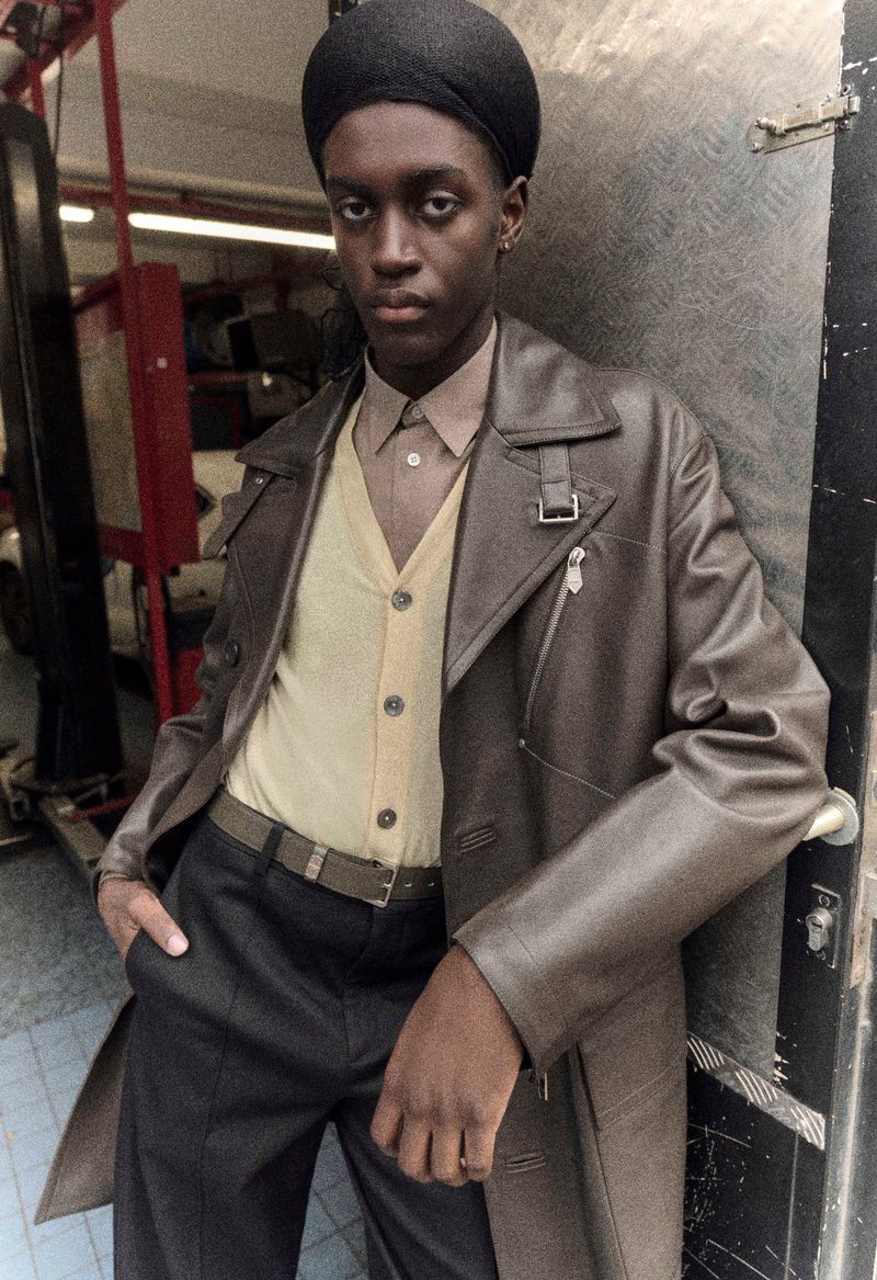 Portrait of a dark-skinned young adult leaning against a doorway, wearing a brown leather trench coat, trousers, and beret in an indoor setting.