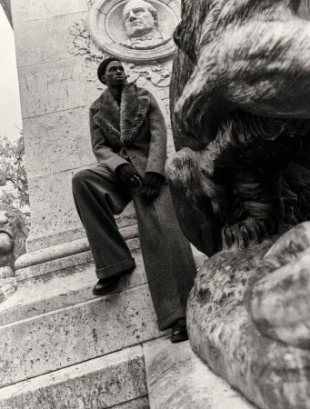 A dark-skinned man in a long coat and hat poses on stone steps next to a lion statue in black and white, looking off to the side.