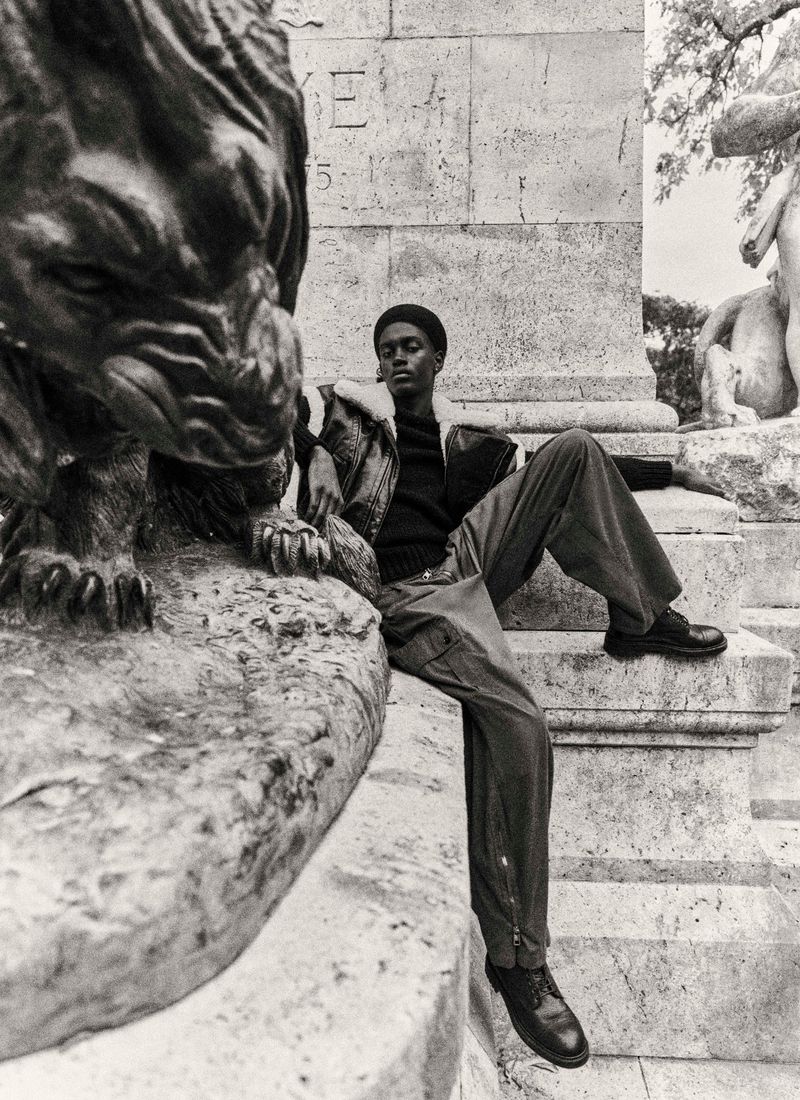 Monochrome fashion shot: Person sits on a stone statue beside a lion statue in an outdoor setting with a stoic expression.
