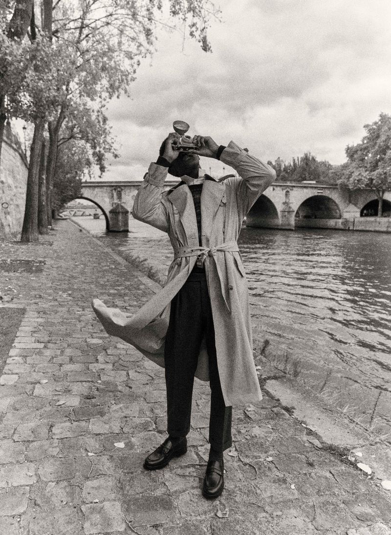 Person in trench coat stands on a cobblestone walkway, holding a vintage camera, with a stone bridge and trees in the background.