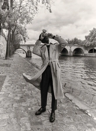 Person in trench coat stands on a cobblestone walkway, holding a vintage camera, with a stone bridge and trees in the background.