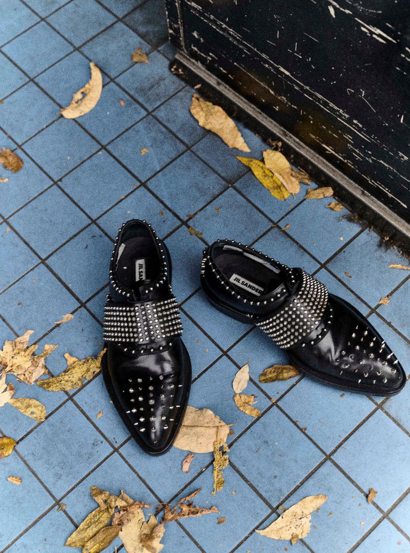 Eye-level shot of two black leather dress shoes with silver studs on a blue tiled floor with scattered yellow and brown leaves.