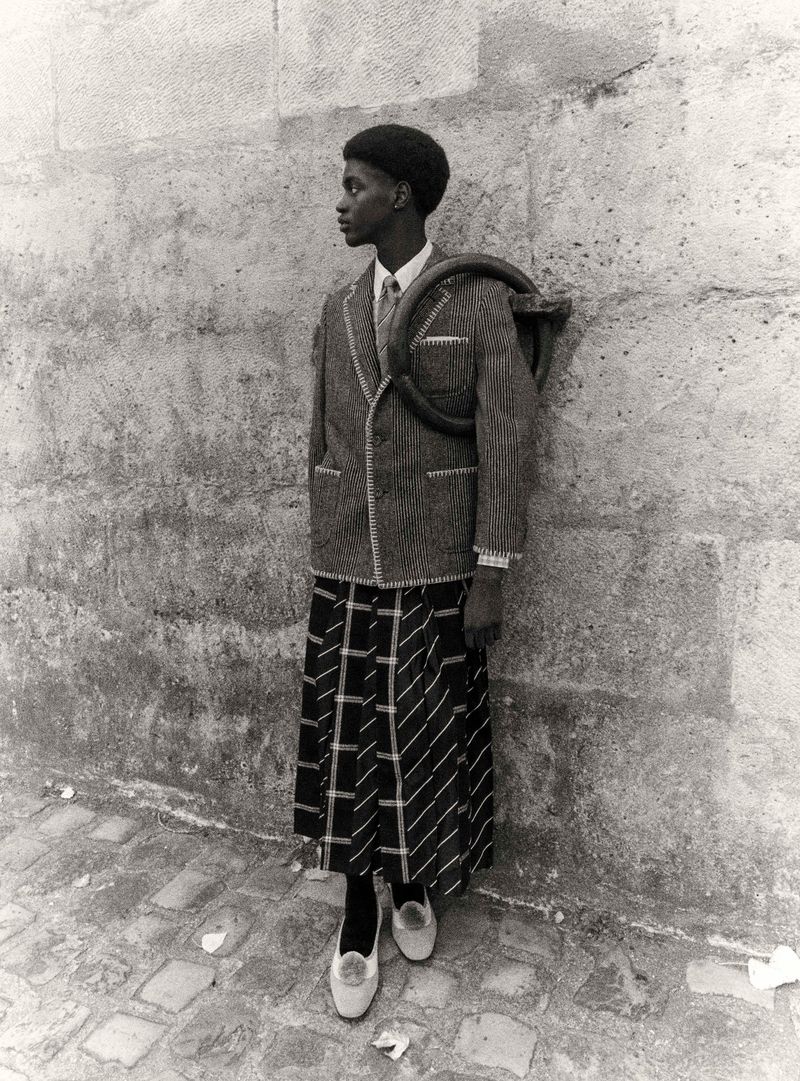Black and white shot of African American person in vintage jacket, skirt, and flats standing against a textured stone wall outdoors.
