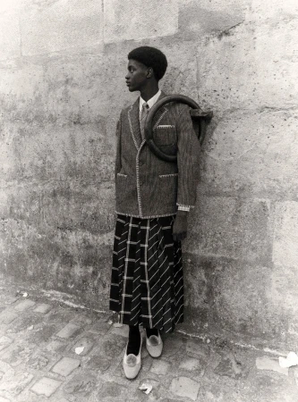 Black and white shot of African American person in vintage jacket, skirt, and flats standing against a textured stone wall outdoors.