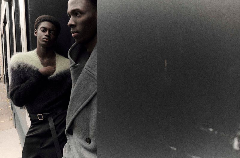 Two dark-skinned men in a fashion portrait. One wears a gray coat, the other a black and white sweater, captured with muted tones.