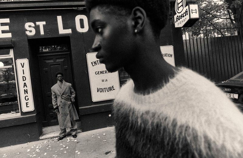 Monochrome portrait of two Black men in an urban setting, one wearing a coat, the other a sweater, with a vintage aesthetic.