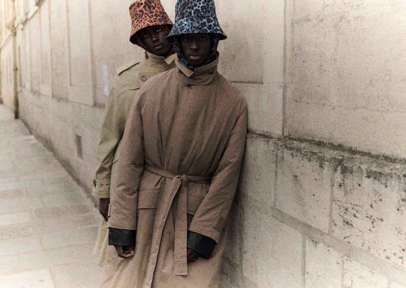 Two adult models in trench coats and leopard-print bucket hats pose against a stone wall, showcasing fashion and urban style.