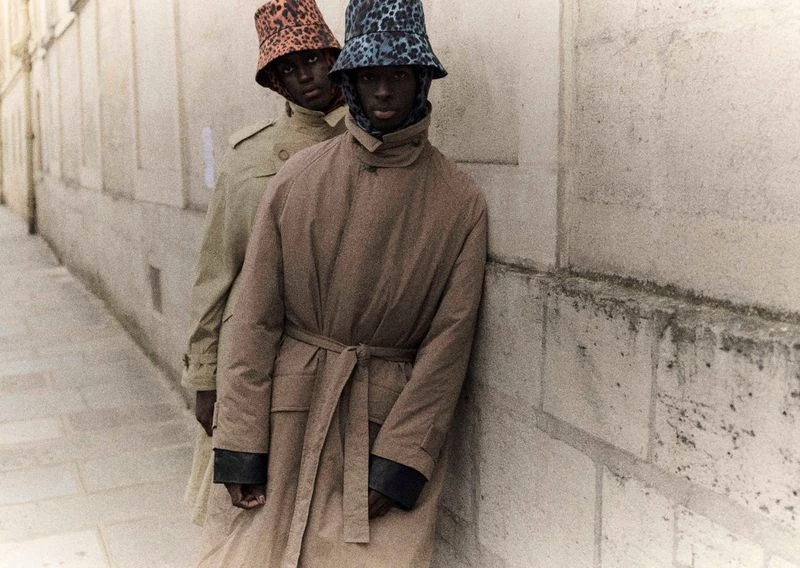 Two adult models in trench coats and leopard-print bucket hats pose against a stone wall, showcasing fashion and urban style.
