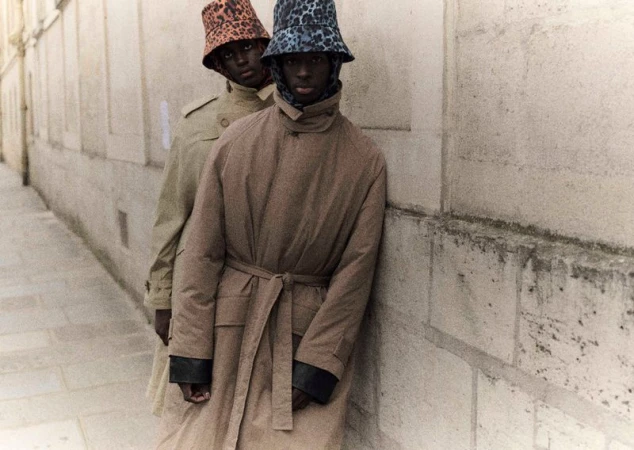 Two adult models in trench coats and leopard-print bucket hats pose against a stone wall, showcasing fashion and urban style.