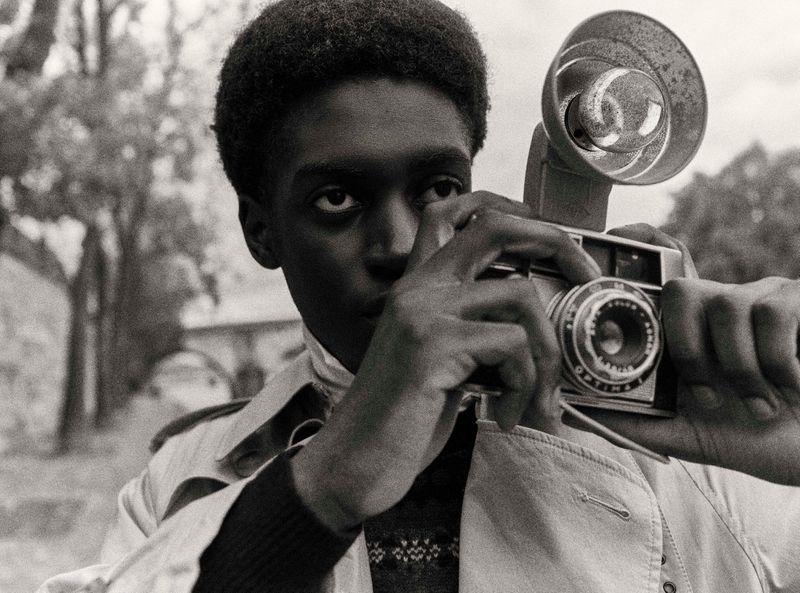 Monochrome image of a young Black man holding a vintage camera with flash, focused expression, outdoors setting with blurred trees.