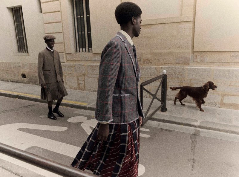 Two African-descent men wearing skirts and suit jackets walk on crosswalk as a brown dog walks behind them along an outdoor city street.