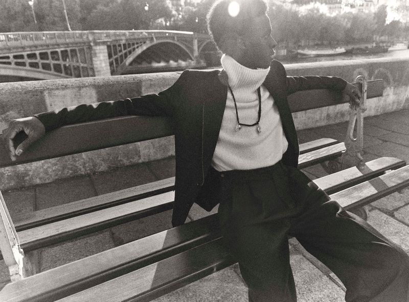 A relaxed, handsome Black man sits on a park bench in a suit and turtleneck, with a bridge visible in the background in a black and white photograph.