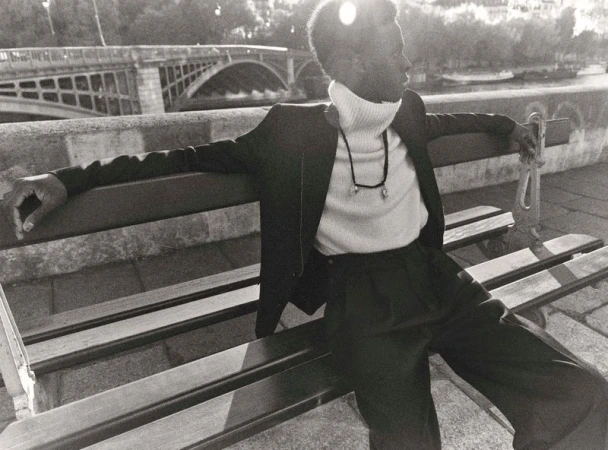 A relaxed, handsome Black man sits on a park bench in a suit and turtleneck, with a bridge visible in the background in a black and white photograph.
