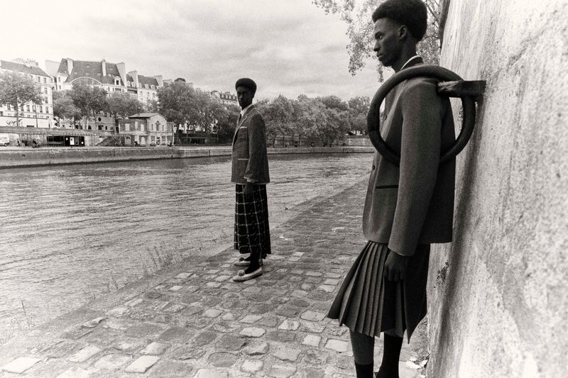 Two African American men in skirts and blazers stand by the Seine River in Paris, showcasing a blend of fashion and architecture.