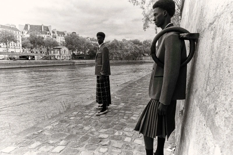 Two African American men in skirts and blazers stand by the Seine River in Paris, showcasing a blend of fashion and architecture.