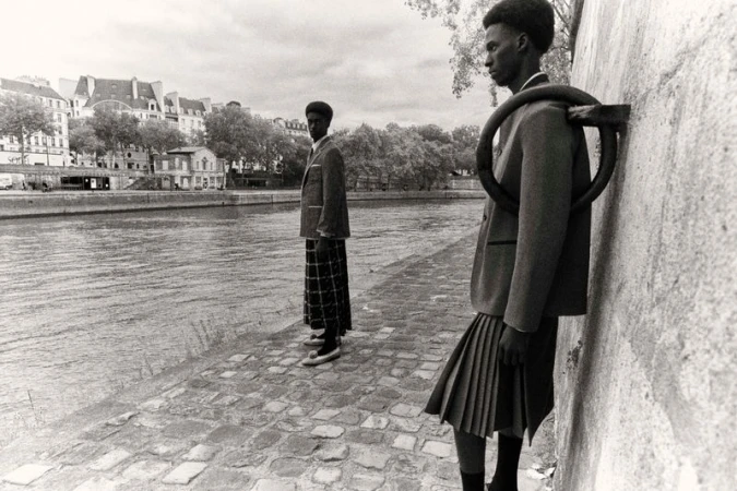 Two African American men in skirts and blazers stand by the Seine River in Paris, showcasing a blend of fashion and architecture.