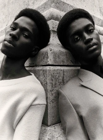 Two Black men lean against a textured stone pillar in a black-and-white fashion portrait wearing a sweater and blazer.