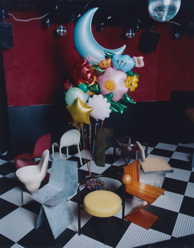 Whimsical balloon bouquet including a crescent moon and flower shapes, arranged among unique chair designs on a checkered floor.