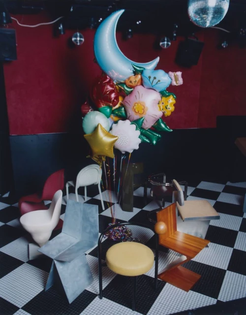 Whimsical balloon bouquet including a crescent moon and flower shapes, arranged among unique chair designs on a checkered floor.