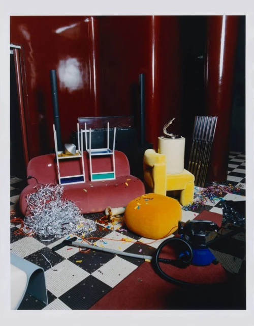 Photo of an indoor scene with a maroon velvet sofa, a yellow chair, various objects, including confetti on a black-and-white checkered floor.