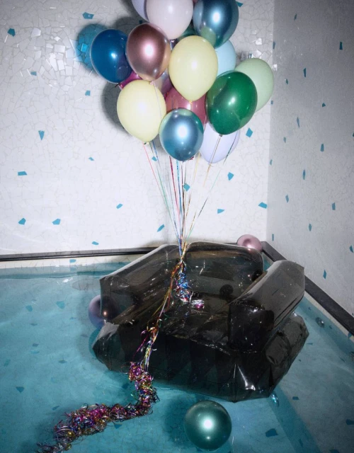 A black inflatable armchair sits in blue water under a cluster of colorful balloons in an indoor setting, creating a whimsical scene.