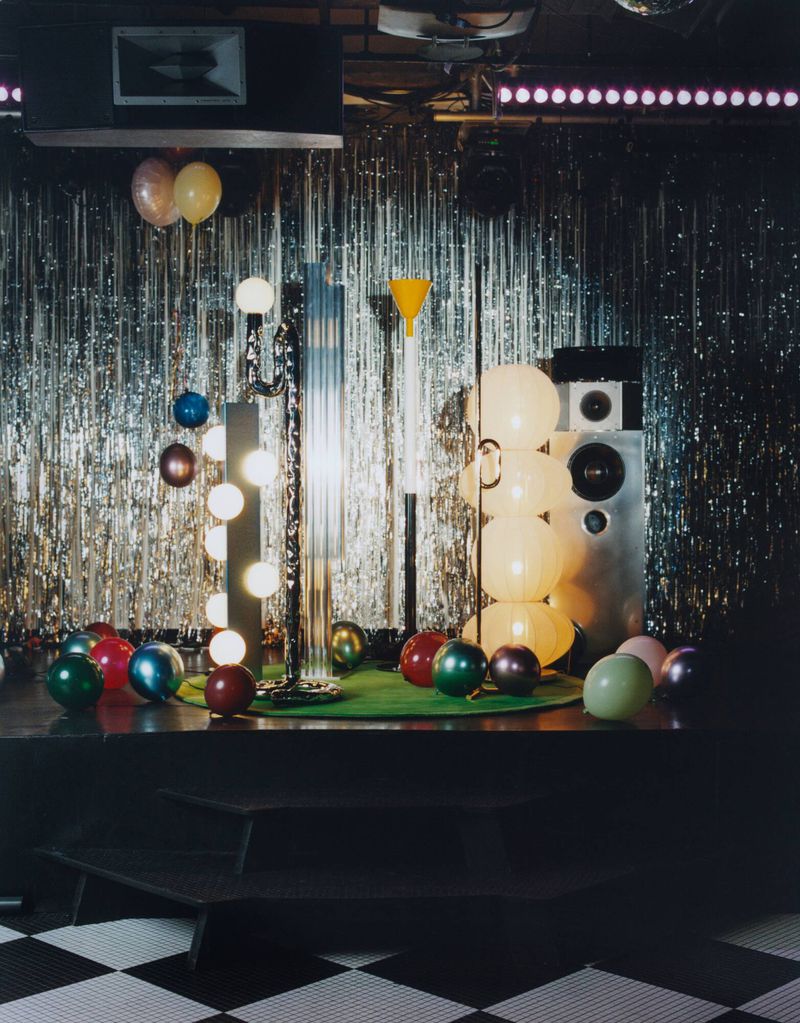 Interior scene featuring disco ball, stage, lighting equipment, and balloons against silver tinsel backdrop, creating a retro party atmosphere.