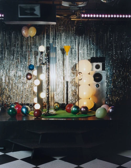Interior scene featuring disco ball, stage, lighting equipment, and balloons against silver tinsel backdrop, creating a retro party atmosphere.