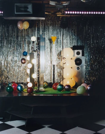 Interior scene featuring disco ball, stage, lighting equipment, and balloons against silver tinsel backdrop, creating a retro party atmosphere.