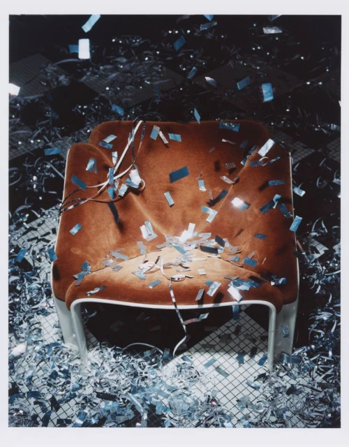 Interior shot of a brown armchair covered in silver confetti, with a matching confetti-covered floor, set against a dark background.