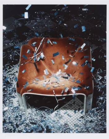 Interior shot of a brown armchair covered in silver confetti, with a matching confetti-covered floor, set against a dark background.