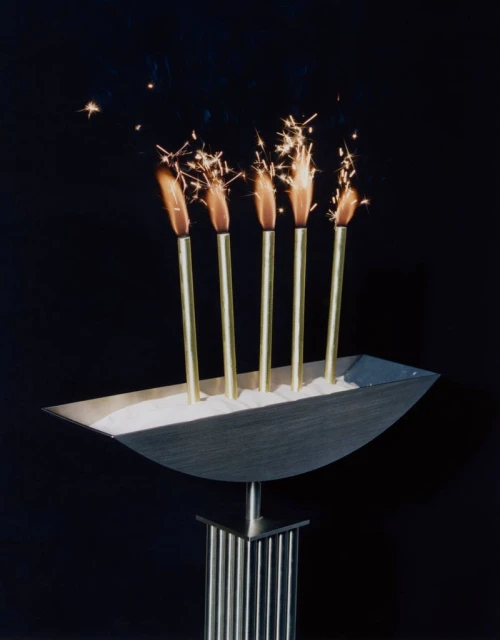 Sparklers burn atop a metallic base filled with white sand against a dark background, creating a festive and celebratory visual.